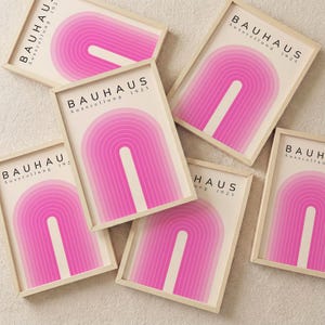 Może przedstawiać: Kilka oprawionych wydruków artystycznych o retro designie. Każdy wydruk przedstawia różowy, łukowaty wzór na kremowym tle, z czarnym napisem "BAUHAUS Ausstellung 1923". Ramki są w jasnym kolorze drewna.