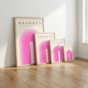 May include: Four framed Bauhaus art prints with a pink arch design. The prints are in varying sizes and feature the text "BAUHAUS Ausstellung 1923" in black. The frames are light-colored wood, and the prints are displayed on a wooden floor.