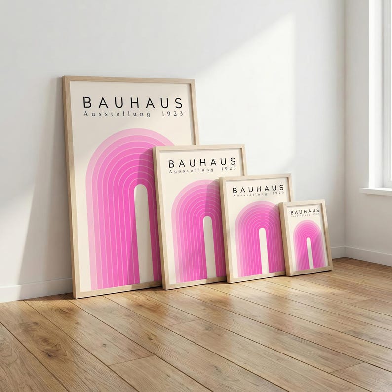 May include: Four framed Bauhaus art prints with a pink arch design. The prints are in varying sizes and feature the text "BAUHAUS Ausstellung 1923" in black. The frames are light-colored wood, and the prints are displayed on a wooden floor.