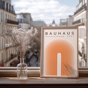 May include: A framed poster for a Bauhaus exhibition, featuring an orange arch design. It's displayed on a wooden windowsill, beside a clear glass vase with dried flowers. The background shows a city street.