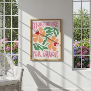 May include: A framed art print with the words "More Amore Por Favor" in pink lettering. The artwork features orange flowers with red centers and green leaves on a cream background. The frame is a light brown color.