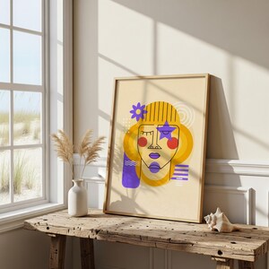 May include: A framed art print featuring a stylized face with yellow hair, a purple star, and a flower. The artwork has a minimalist design with bold lines and a neutral color palette. The print is displayed on a rustic wooden table.