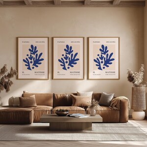 May include: Three framed art prints with blue abstract floral designs are displayed above a brown sectional sofa. The prints have a beige background and are labeled "PAPIERS DÉCOUPÉS MATISSE". A wooden coffee table and a rug complete the living room.