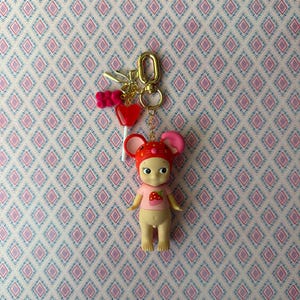 Puede incluir: Un llavero con una peque&ntilde;a mu&ntilde;eca con un atuendo con tem&aacute;tica de fresa y un sombrero rojo con orejas rosas. La mu&ntilde;eca est&aacute; unida a un llavero dorado con dijes, incluido un coraz&oacute;n rojo y un lazo rosa. El fondo es un papel pintado estampado.