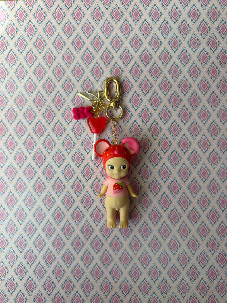 Puede incluir: Un llavero con una peque&ntilde;a mu&ntilde;eca con un atuendo con tem&aacute;tica de fresa y un sombrero rojo con orejas rosas. La mu&ntilde;eca est&aacute; unida a un llavero dorado con dijes, incluido un coraz&oacute;n rojo y un lazo rosa. El fondo es un papel pintado estampado.