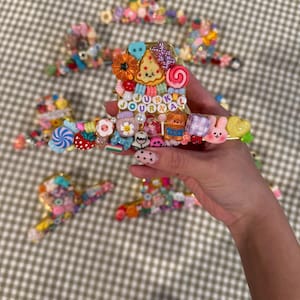 May include: A gold-toned hair claw clip decorated with an assortment of colorful miniature charms including a pizza slice, a teddy bear, flowers, candies, and beads spelling out JUNK JOURNAL, surrounded by several similar decorative hair clips.