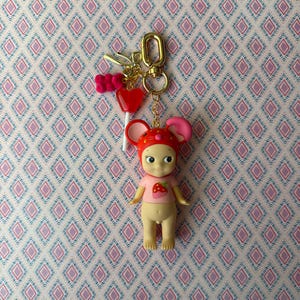 Puede incluir: Un llavero caprichoso con una peque&ntilde;a mu&ntilde;eca con gorro de seta rojo y una camisa rosa con una fresa. El llavero incluye un cierre dorado, una piruleta de coraz&oacute;n rojo y un charm de osito de goma rosa.