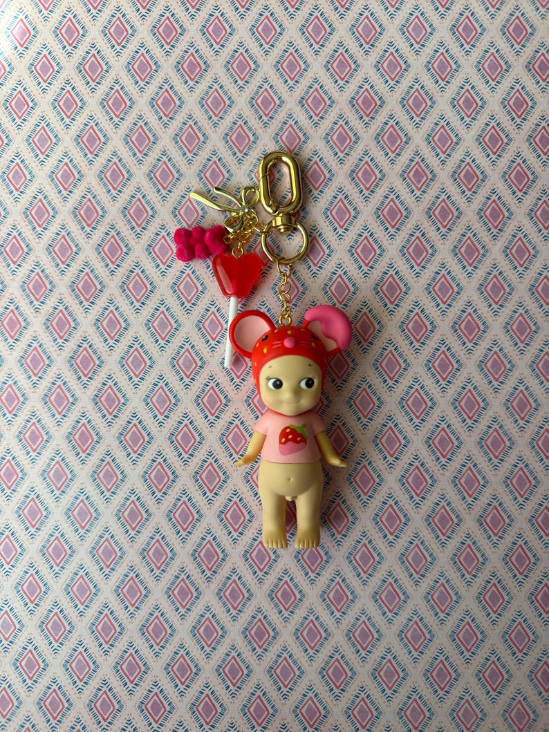 Puede incluir: Un llavero caprichoso con una peque&ntilde;a mu&ntilde;eca con gorro de seta rojo y una camisa rosa con una fresa. El llavero incluye un cierre dorado, una piruleta de coraz&oacute;n rojo y un charm de osito de goma rosa.