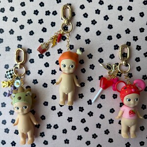 Puede incluir: Tres peque&ntilde;as figuras de color nude con llaveros. Cada figura tiene un sombrero y un amuleto diferentes. Una tiene un sombrero de oso, otra un sombrero naranja y la &uacute;ltima un sombrero de fresa. Los amuletos incluyen un rayo y una taza peque&ntilde;a.