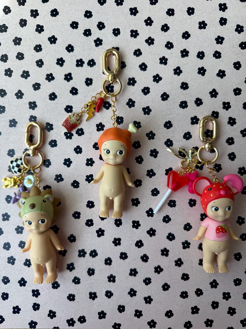 Puede incluir: Tres peque&ntilde;as figuras de color nude con llaveros. Cada figura tiene un sombrero y un amuleto diferentes. Una tiene un sombrero de oso, otra un sombrero naranja y la &uacute;ltima un sombrero de fresa. Los amuletos incluyen un rayo y una taza peque&ntilde;a.