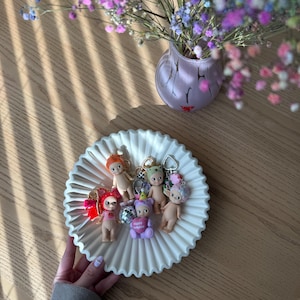 Puede incluir: Un plato de cer&aacute;mica blanca estriada contiene varios llaveros de mu&ntilde;ecas peque&ntilde;as. Los llaveros presentan varios dise&ntilde;os de mu&ntilde;ecas con diferentes accesorios. Un jarr&oacute;n de flores coloridas se encuentra cerca sobre una mesa de madera.