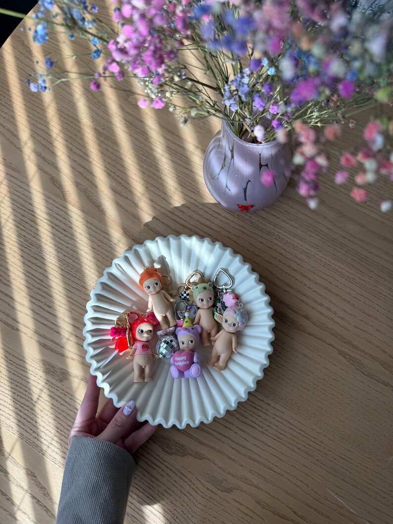 Puede incluir: Un plato de cer&aacute;mica blanca estriada contiene varios llaveros de mu&ntilde;ecas peque&ntilde;as. Los llaveros presentan varios dise&ntilde;os de mu&ntilde;ecas con diferentes accesorios. Un jarr&oacute;n de flores coloridas se encuentra cerca sobre una mesa de madera.