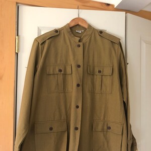 May include: Olive green jacket with a mandarin collar, button-down front, and four front pockets. The jacket has shoulder epaulets and long sleeves. It is hanging on a wooden hanger against a white door.