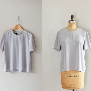 May include: A light gray, short-sleeved blouse with a round neckline. The blouse is made of a smooth, silky fabric.
