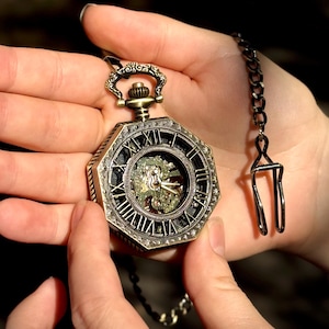 Może przedstawiać: Antyczny zegarek kieszonkowy z brązową obudową i łańcuszkiem. Zegarek ma szczegółową tarczę z rzymskimi cyframi i widocznym mechanizmem wewnętrznym. Zegarek jest trzymany w otwartych dłoniach na rozmytym tle.