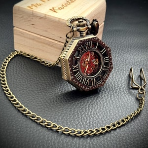 May include: An octagonal bronze-toned pocket watch with a red skeleton dial and Roman numerals, attached to a long metal chain with a belt clip. The watch rests in front of a light-colored wooden box with the text Mr. Maximos Karattas engraved on top.