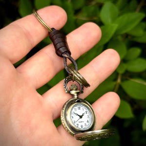 May include: A bronze-coloured pocket watch with a white clock face and black numerals. The watch is open, revealing the time. It is attached to a leather strap and a key ring. The watch case has decorative detailing.