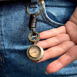 May include: A vintage-style pocket watch keychain with a bronze-colored case and a cream-colored watch face. The keychain features a brown leather strap and a brass-colored ring. The watch is attached to a brass-colored ring that is attached to a brown leather strap.