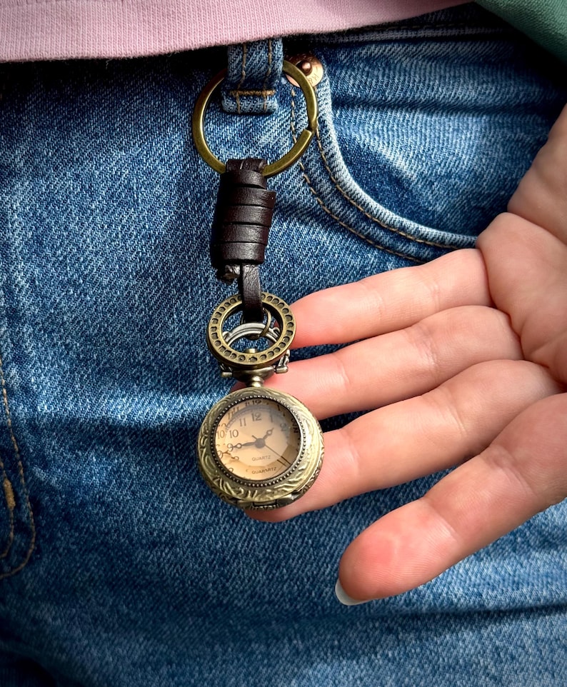 May include: A vintage-style pocket watch keychain with a bronze-colored case and a cream-colored watch face. The keychain features a brown leather strap and a brass-colored ring. The watch is attached to a brass-colored ring that is attached to a brown leather strap.