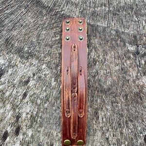 May include: A brown leather cuff bracelet with brass-colored rivets and fasteners. The cuff has a distressed finish and a series of adjustable straps. The bracelet is designed to wrap around the wrist.