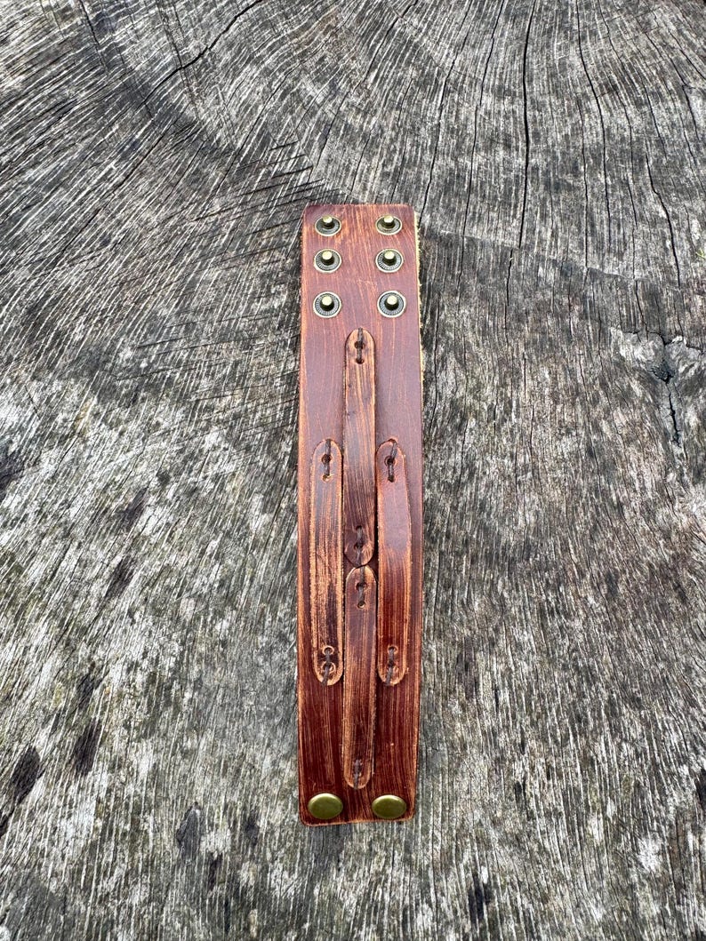 May include: A brown leather cuff bracelet with brass-colored rivets and fasteners. The cuff has a distressed finish and a series of adjustable straps. The bracelet is designed to wrap around the wrist.