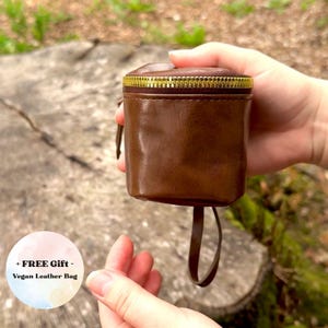 May include: A brown vegan leather bag with a gold zipper closure. The small, cylindrical bag has a drawstring and is held in front of a natural background. The text "+ FREE Gift - Vegan Leather Bag" is in a circle in the lower left.