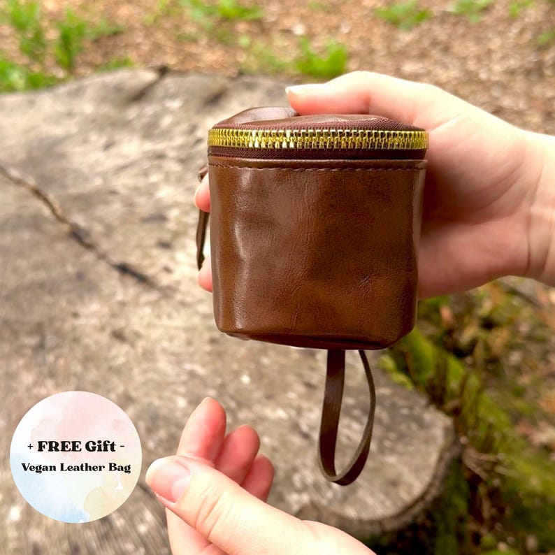 May include: A brown vegan leather bag with a gold zipper closure. The small, cylindrical bag has a drawstring and is held in front of a natural background. The text "+ FREE Gift - Vegan Leather Bag" is in a circle in the lower left.