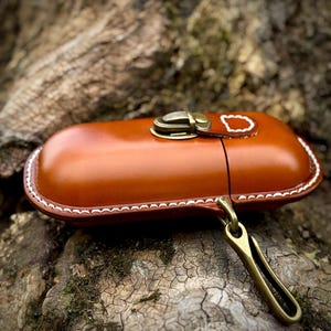 May include: A tan leather case with a brass clasp and a brass hook. The case has white stitching around the edges. The case is resting on a textured, brown surface.