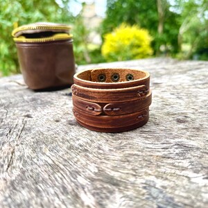 May include: A stack of brown leather bracelets with decorative stitching and metal rivets. A small, brown leather pouch with a gold zipper is in the background. The items are on a weathered wooden surface.
