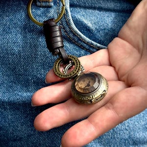 May include: A vintage-style pocket watch with a detailed bronze case and a clear, faceted crystal. The watch face displays Roman numerals. It is attached to a key ring with a brown leather strap and bronze hardware. The watch is held against a denim background.