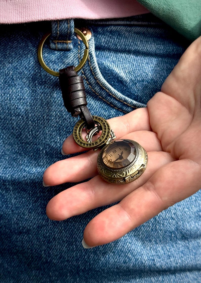 May include: A vintage-style pocket watch with a detailed bronze case and a clear, faceted crystal. The watch face displays Roman numerals. It is attached to a key ring with a brown leather strap and bronze hardware. The watch is held against a denim background.