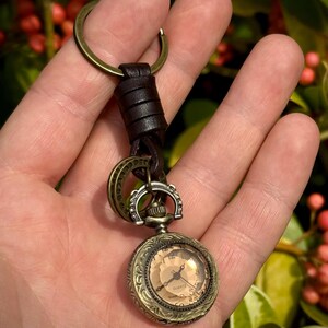 May include: A vintage-style pocket watch keychain with a bronze-colored case and a cream-colored face. The watch is attached to a leather strap and a bronze key ring. The background is blurred, with green foliage and red berries.