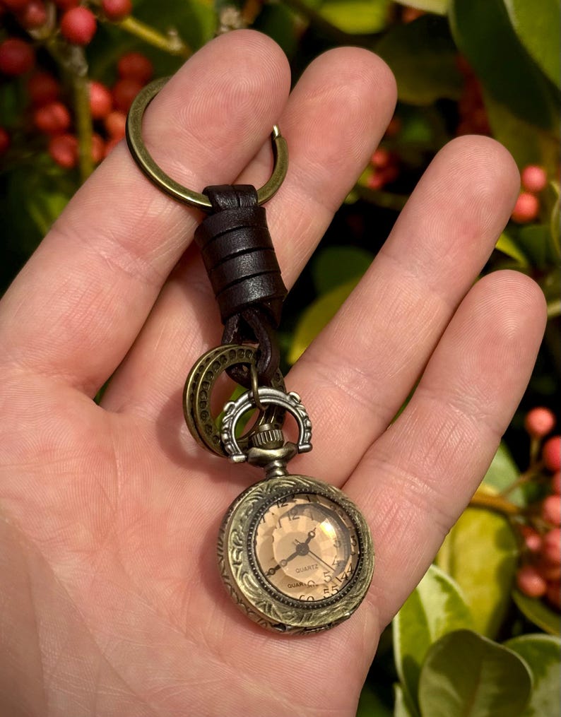 May include: A vintage-style pocket watch keychain with a bronze-colored case and a cream-colored face. The watch is attached to a leather strap and a bronze key ring. The background is blurred, with green foliage and red berries.