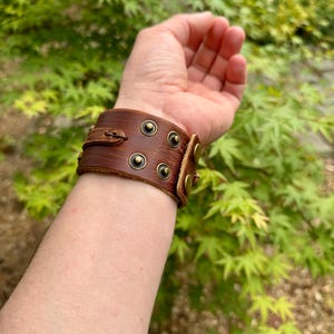May include: A brown leather cuff bracelet with brass-colored rivets and a buckle closure. The bracelet is worn on a wrist, showcasing its detailed design and craftsmanship. The background features green foliage.