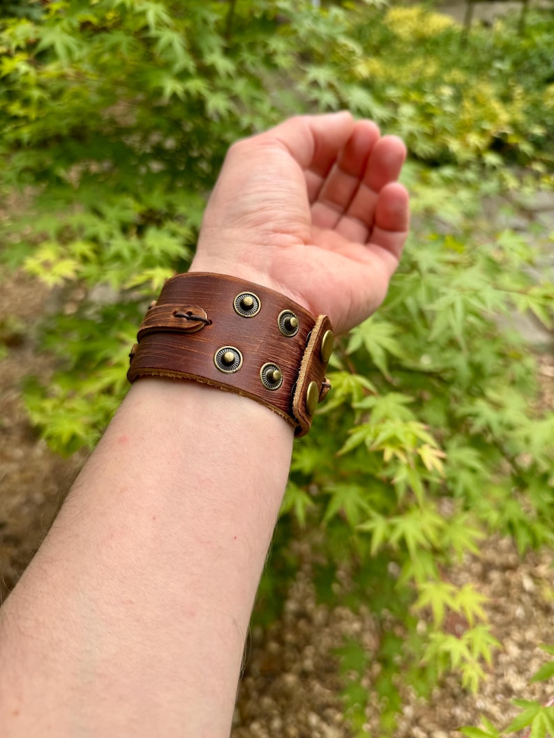 May include: A brown leather cuff bracelet with brass-colored rivets and a buckle closure. The bracelet is worn on a wrist, showcasing its detailed design and craftsmanship. The background features green foliage.