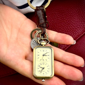 Puede incluir: Un llavero de estilo vintage con un reloj rectangular dorado con dos esferas. El reloj está sujeto a una correa de cuero marrón y un llavero de latón. Cuelgan dijes decorativos adicionales debajo del reloj.