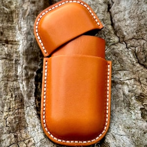 May include: A handcrafted, open leather case in a warm, orange-brown hue. The case features a rounded top and bottom with white stitching along the edges. The top flap is open, revealing the interior. The case is likely designed to hold a small item.