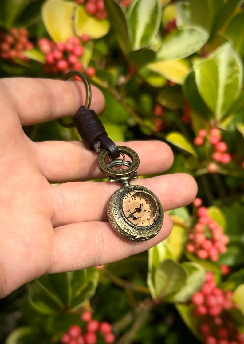 May include: Antique bronze-colored pocket watch keychain with a brown leather strap. The watch has a detailed face with black hands and Roman numerals. The keychain is held against a backdrop of green foliage and red berries.