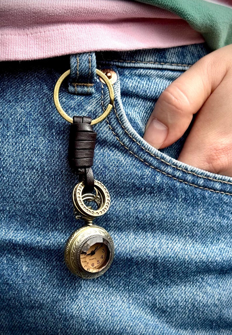 May include: A vintage-style pocket watch keychain with a bronze finish. The watch features a small, round face with Roman numerals and a quartz movement. The keychain includes a leather strap and a brass ring, attached to a pair of blue denim jeans.