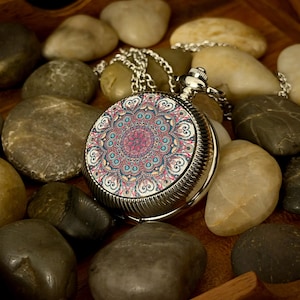 May include: A silver pocket watch with a decorative, colourful mandala design on the face. The watch is on a silver chain and rests among smooth, grey and tan stones. The watch face features pink, blue, and yellow patterns.