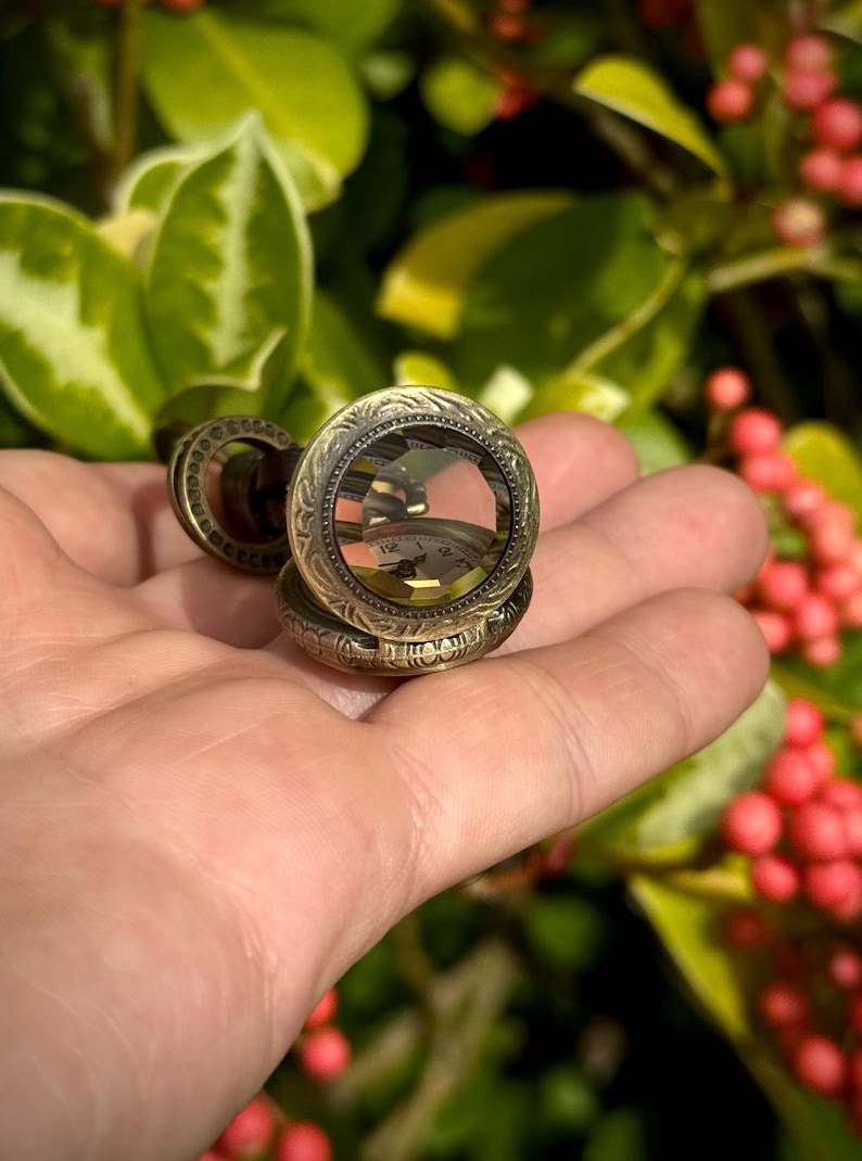 May include: An antique-style pocket watch with an ornate, engraved design. The watch has a clear crystal face, revealing the clock hands and numbers. The watch is a bronze color and appears to be made of metal.