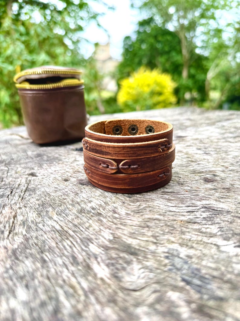 May include: A stack of brown leather bracelets with decorative stitching and metal rivets. A small, brown leather pouch with a gold zipper is in the background. The items are on a weathered wooden surface.