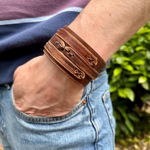 May include: Wide brown leather cuff bracelet with three decorative buttons. The bracelet is worn on a wrist, and the person is wearing a blue and purple striped shirt and blue jeans.