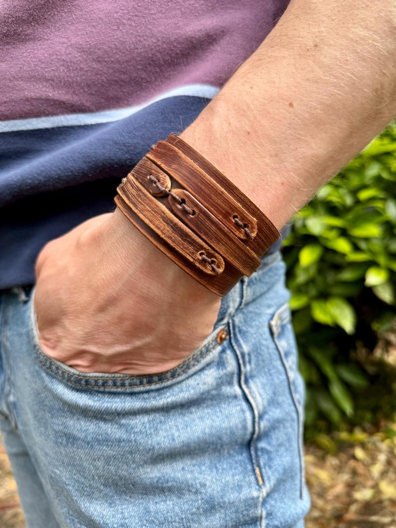 May include: Wide brown leather cuff bracelet with three decorative buttons. The bracelet is worn on a wrist, and the person is wearing a blue and purple striped shirt and blue jeans.