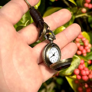 May include: A vintage-style pocket watch with an open face, revealing a white clock face with black numerals and hands. The watch has an antique brass finish and is attached to a brown leather strap. The watch is held in a hand against a blurred green and red background.