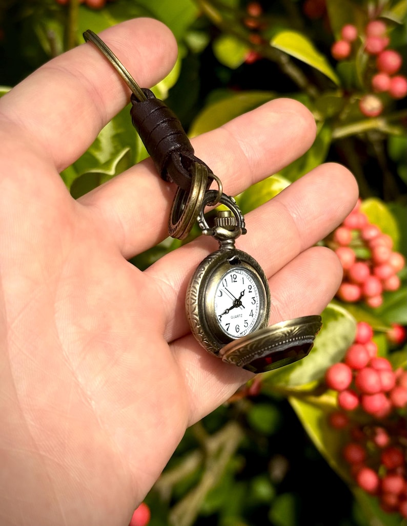 May include: A vintage-style pocket watch with an open face, revealing a white clock face with black numerals and hands. The watch has an antique brass finish and is attached to a brown leather strap. The watch is held in a hand against a blurred green and red background.