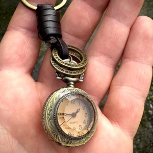 May include: Antique-style pocket watch keychain with a bronze finish. The watch face is visible, displaying the time. The keychain includes a leather strap and bronze rings for attachment. The watch reads "QUARTZ".
