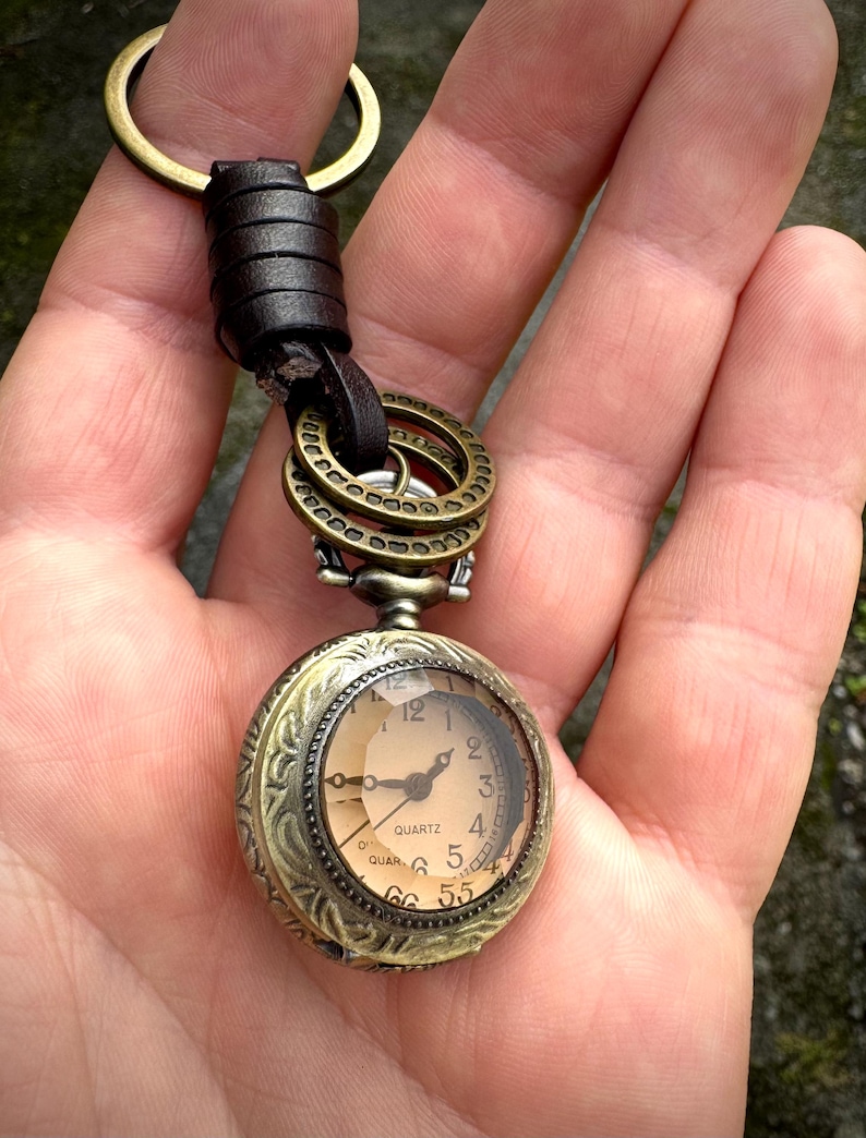 May include: Antique-style pocket watch keychain with a bronze finish. The watch face is visible, displaying the time. The keychain includes a leather strap and bronze rings for attachment. The watch reads "QUARTZ".