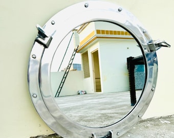 May include: A circular polished silver-tone metal porthole mirror featuring a wide frame with evenly spaced rivets, two side-mounted locking handles, and a bottom hinge mechanism, reflecting an outdoor building scene.