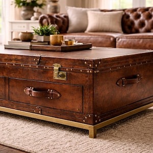 Op de afbeelding: Een rechthoekige bruin lederen koffietafel in kofferstijl met messing accenten. De tafel heeft twee lades met leren handgrepen en een messing slot. De tafel staat op een messing frame.